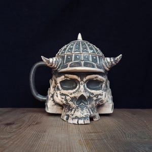 Könnte beinhalten: Ein Keramikbecher in Form eines menschlichen Schädels mit gehörntem Helm. Der Becher hat ein verwittertes grau-weißes Finish mit dunklen Details in den Vertiefungen. Ein geschwungener Henkel ragt seitlich heraus und der Helm hat eine Spitze.