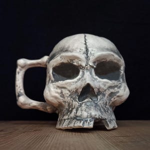 Może przedstawiać: Ceramiczny kubek w kształcie ludzkiej czaszki o postarzanym, złamanej bieli i szarym wykończeniu. Uchwyt zaprojektowano tak, aby przypominał kość, a czaszka ma głębokie, ciemne oczodoły i jamę nosową oraz widoczny szew na sklepieniu czaszki.