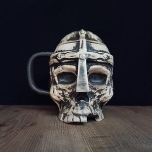 Peut inclure: Une tasse en céramique en forme de crâne humain portant un casque de style métallique. La tasse présente une finition vieillie blanc cassé et gris foncé avec une crête en forme de croix sur le front et une anse incurvée foncée sur le côté gauche.
