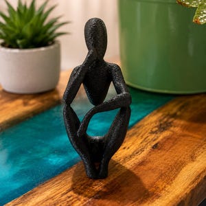Op de afbeelding: Een zwart, getextureerd, abstract menselijk beeldje in een zittende, peinzende houding met één hand bij het gezicht. Het beeldhouwwerk heeft een ruwe, korrelige oppervlakteafwerking en een slanke, gestileerde vorm die rust uitstraalt.