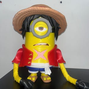 Può includere: Una statuetta gialla di un personaggio Minion che indossa un cappello di paglia, un gilet rosso aperto con bottoni dorati, pantaloncini blu e una fascia bianca. La figura ha un occhio solo con occhiali grigi, capelli neri e una cicatrice rosa a forma di X sul petto giallo.