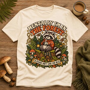 Könnte beinhalten: Ein cremefarbenes T-Shirt mit einer Grafik eines Waschbären in einem roten Pullover, der auf einem Baumstumpf sitzt und eine Tasse hält. Der Text lautet MENTALLY I'M IN THE FOREST und PLEASE DON'T LOOK FOR ME, umgeben von Bäumen, Pilzen und einer Laterne.