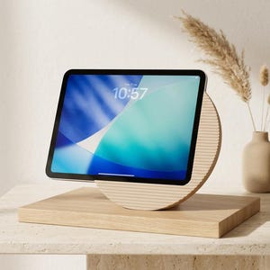 Może przedstawiać: Tablet z niebiesko-białym ekranem wyświetlającym godzinę 10:57, umieszczony na jasnym drewnianym stojaku z okrągłą podstawą z poziomymi rowkami oraz płaską prostokątną platformą zapewniającą stabilne podparcie urządzenia.