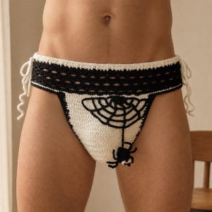 Puede incluir: Una braguita de bikini de ganchillo en blanco y negro con un motivo de telaraña y una pequeña aplicación de araña negra. La prenda tiene una cintura ancha negra con un patrón calado decorativo y lazos laterales blancos para ajustar.