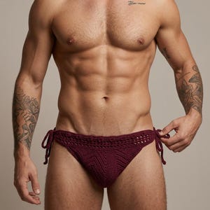 Puede incluir: Una braguita de bikini de ganchillo en color burdeos oscuro que presenta un patrón de chevrón texturizado en el panel frontal, una cintura decorativa ancha con diseño de punto abierto y lazos laterales ajustables anudados a ambos lados.