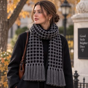 May include: A long, rectangular crochet scarf featuring a grid pattern of black squares outlined by thick, textured gray yarn. The scarf ends are finished with long, straight gray fringe tassels. The scarf is worn over a black coat and a brown bag.