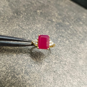 May include: A gold ring featuring a large, rectangular, vibrant pinkish-red gemstone set in the center. The band is accented with three small, clear, round-cut stones on each side of the central setting, creating a balanced and elegant appearance.