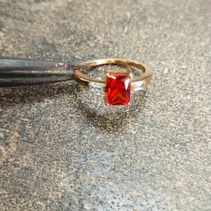 May include: A gold-toned ring featuring a central rectangular-cut vibrant red gemstone flanked by two smaller clear baguette-cut stones on either side of the band. The ring is held by a pair of metal tweezers, showcasing the setting and stone clarity.