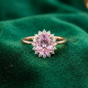 Puede incluir: Un anillo de oro rosa con una piedra preciosa rosa de corte ovalado en el centro, rodeada por un halo de pequeñas piedras blancas redondas y transparentes. El aro es liso y pulido, con un engaste de garras clásico para las piedras.