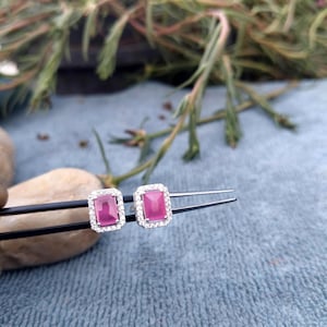 Puede incluir: Un par de pendientes de botón con piedras centrales rosas rectangulares rodeadas por un halo de pequeñas piedras blancas transparentes. Los pendientes se sujetan con unas pinzas negras, mostrando los engastes plateados y el color rosa.