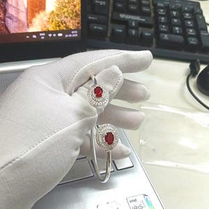 Puede incluir: Una pulsera abierta de tono plateado con dos piedras rojas de forma ovalada en cada extremo. Cada piedra roja está rodeada por un doble halo de pequeñas piedras claras y brillantes engastadas en un marco circular, creando un diseño simétrico.