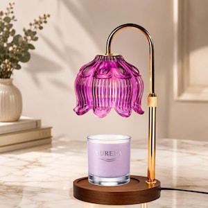 Puede incluir: Una lámpara calentadora de velas con soporte de metal dorado de brazo curvo y base de madera oscura. Una pantalla de vidrio morado con forma de flor festoneada cuelga sobre un frasco de vela transparente con cera morada clara y el texto LUREXA.