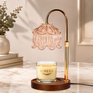 Può includere: Una lampada scaldacandela con supporto in metallo dorato, base circolare in legno scuro e paralume in vetro scanalato rosa pallido a forma di fiore. Una candela in barattolo di vetro trasparente con cera color crema e scritta dorata LUREXA.
