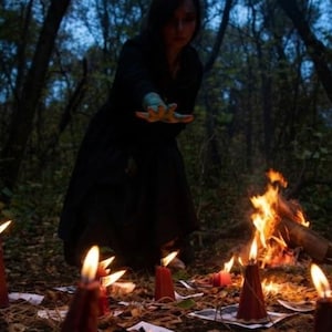 May include: A person in a black hooded garment reaches out with a hand over a collection of red pillar candles arranged on the ground. Each candle sits atop a small piece of paper. A small campfire burns in the background of the wooded area.