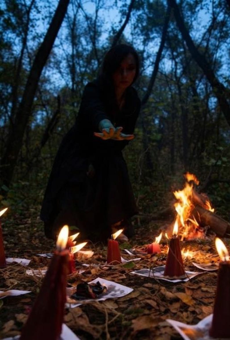 May include: A person in a black hooded garment reaches out with a hand over a collection of red pillar candles arranged on the ground. Each candle sits atop a small piece of paper. A small campfire burns in the background of the wooded area.