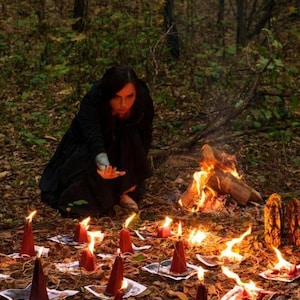 May include: A collection of dark red, cone-shaped candles arranged in a circular pattern on a forest floor covered in fallen leaves. Each candle sits atop a small square paper print, with flames flickering above the wicks in a dim outdoor setting.