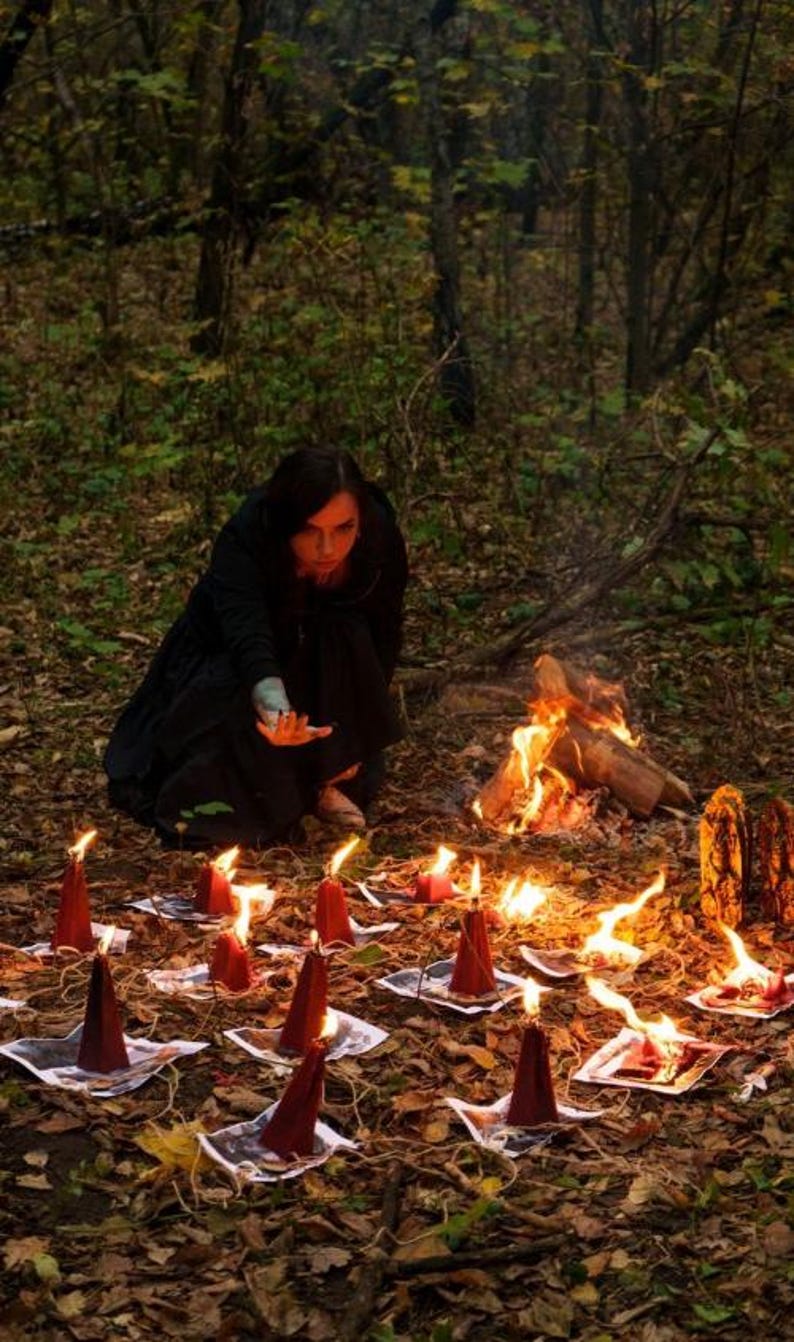May include: A collection of dark red, cone-shaped candles arranged in a circular pattern on a forest floor covered in fallen leaves. Each candle sits atop a small square paper print, with flames flickering above the wicks in a dim outdoor setting.