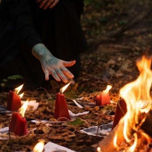 Puede incluir: Una colecci&oacute;n de velas rojas en forma de cono dispuestas en c&iacute;rculo sobre el suelo, cada una colocada sobre un peque&ntilde;o cuadrado de papel blanco. Las velas est&aacute;n encendidas, produciendo llamas brillantes, mientras un fuego mayor arde cerca.