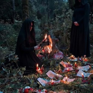May include: Two figures in black hooded cloaks stand in a forest clearing. One kneels by a small fire, while the other stands nearby. Scattered on the ground are burning papers, small animal figurines, pomegranates, nuts, and floral elements.