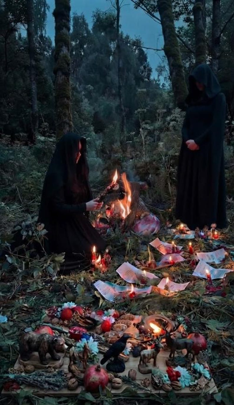 May include: Two figures in black hooded cloaks stand in a forest clearing. One kneels by a small fire, while the other stands nearby. Scattered on the ground are burning papers, small animal figurines, pomegranates, nuts, and floral elements.