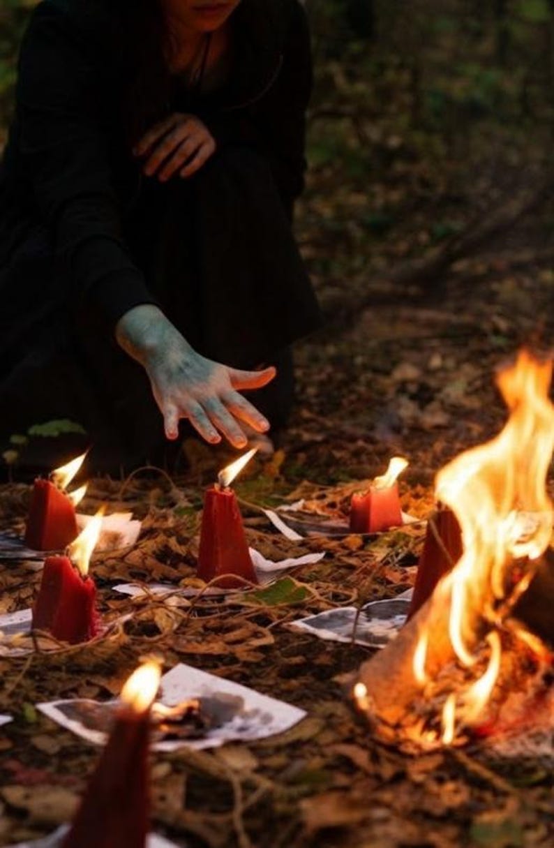 May include: A collection of red conical candles arranged on the ground, each placed on a small white paper square. The candles are lit, casting a warm glow, while a larger fire burns nearby. The scene is set on a bed of dry autumn leaves.