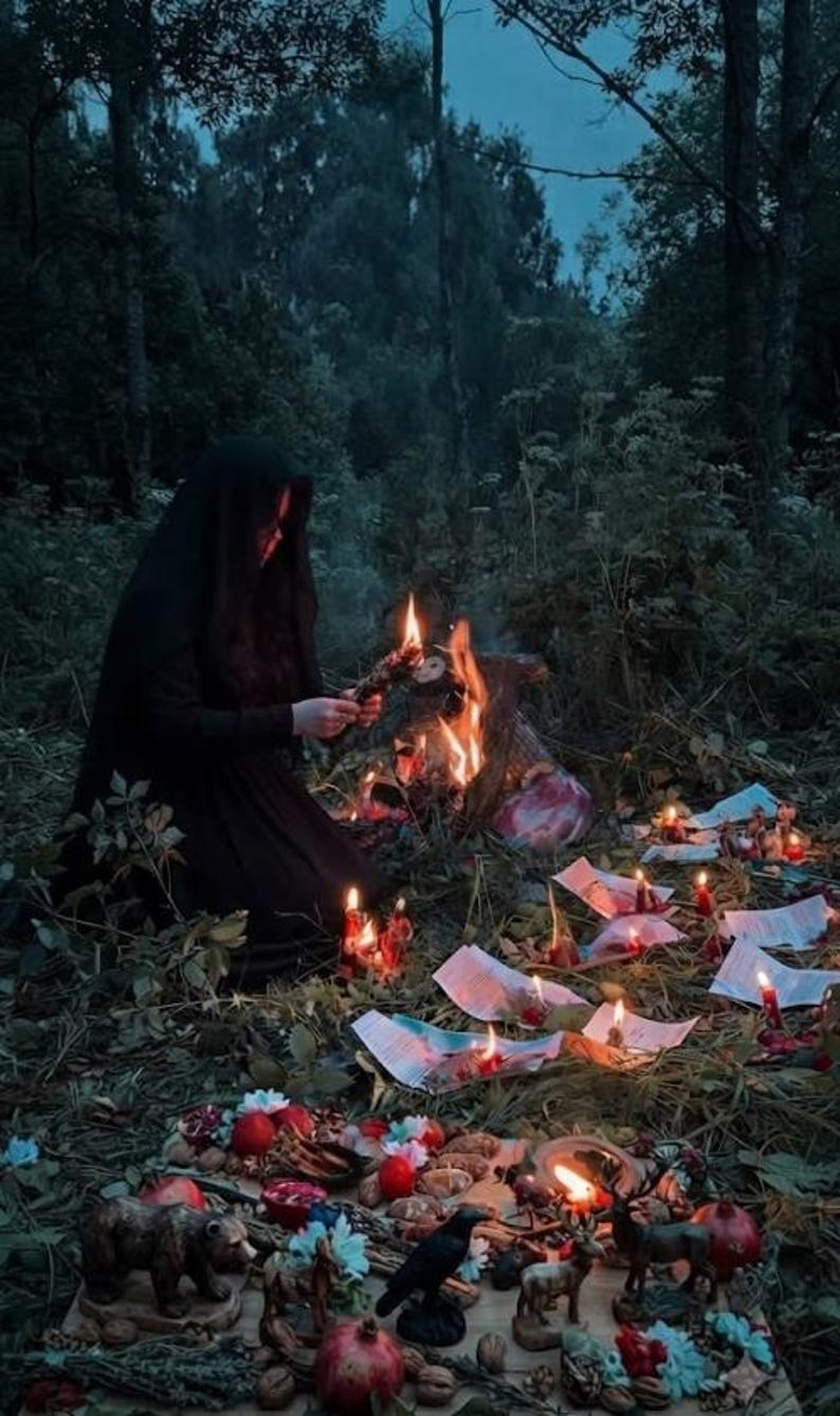 Puede incluir: Una persona con una capa negra con capucha arrodillada junto a una hoguera en un bosque, rodeada de papeles esparcidos, velas encendidas y una colecci&oacute;n de peque&ntilde;as figuras de animales como un oso, un cuervo y un ciervo, junto a granadas y nueces.