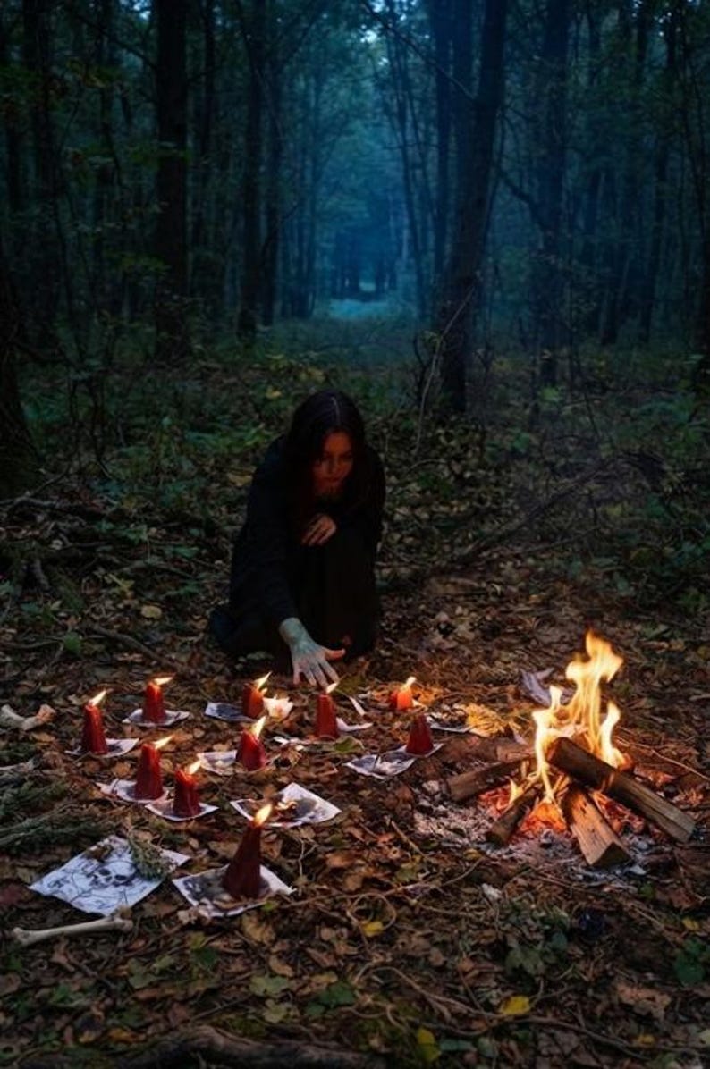 May include: A person in dark clothing kneels in a forest clearing near a small campfire and a circle of red pillar candles placed on top of white paper sheets featuring skull illustrations, with scattered bones on the ground nearby.