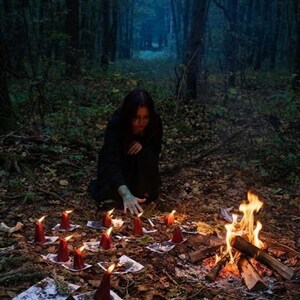 Puede incluir: Una persona con ropa oscura arrodillada en un bosque cerca de una peque&ntilde;a fogata y un c&iacute;rculo de velas de pilar rojas sobre hojas de papel blanco, algunas con ilustraciones de calaveras, entre hojas secas y fragmentos de huesos.