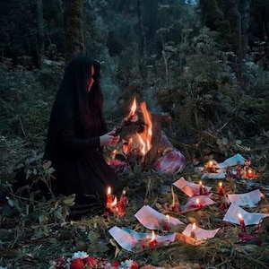 May include: A person in a black hooded cloak kneels by a fire in a forest. Scattered papers, candles, and a wooden board featuring small animal figurines, pomegranates, nuts, and floral accents are arranged on the ground in front of the fire.