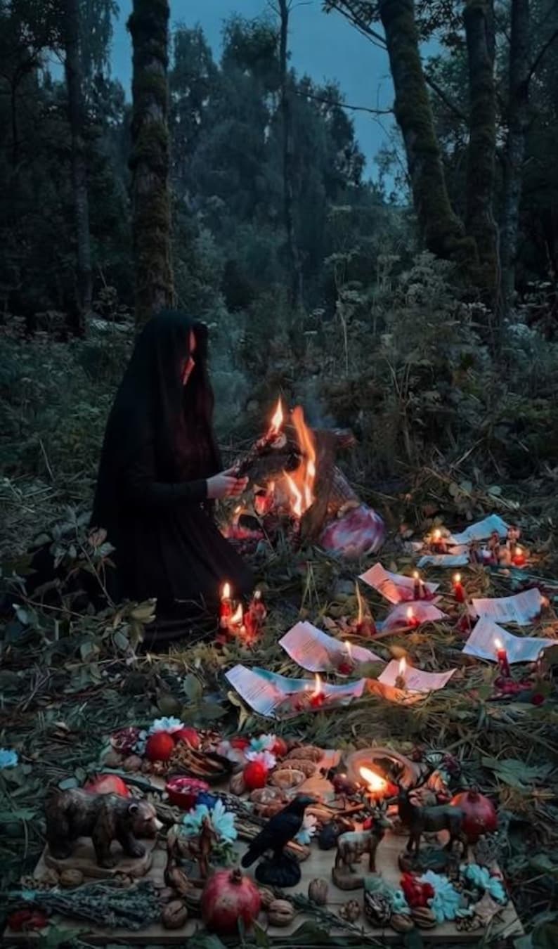 May include: A person in a black hooded cloak kneels by a fire in a forest. Scattered papers, candles, and a wooden board featuring small animal figurines, pomegranates, nuts, and floral accents are arranged on the ground in front of the fire.