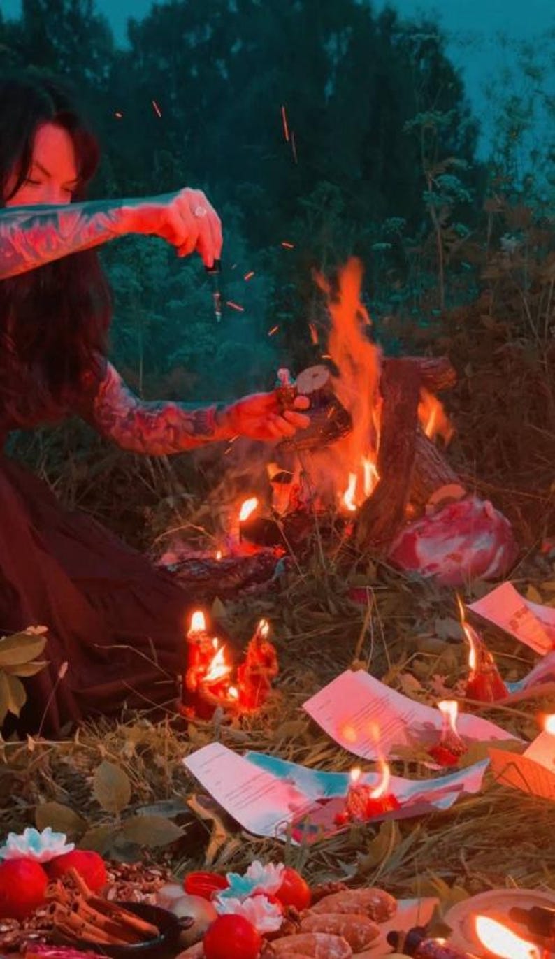 May include: A glass dropper bottle held over a small campfire, surrounded by scattered papers with printed text, red candles, cinnamon sticks, and small white flowers on the ground, illuminated by the orange glow of the flames.