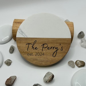 May include: A circular coaster featuring a split design with white faux marble on the top half and natural wood grain on the bottom half. The wood section is engraved with the black text "The Perry's" and "est. 2024" in a script and sans-serif font.