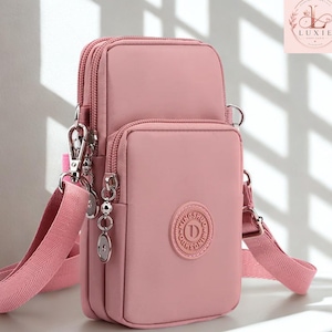 May include: A dusty pink crossbody bag featuring two zippered compartments, a detachable adjustable shoulder strap, and a circular emblem on the front with the text MINGSHUO. The bag is constructed from a smooth, solid-colored fabric material.