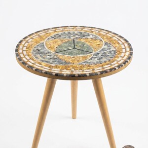 Op de afbeelding: Een ronde bijzettafel met drie lichtgekleurde houten poten en een rond tafelblad met een mozaïekpatroon. Het mozaïek bestaat uit kleine vierkante tegeltjes in tinten geel, grijs, wit en groen, gerangschikt in een geometrisch ontwerp van in elkaar grijpende cirkels.
