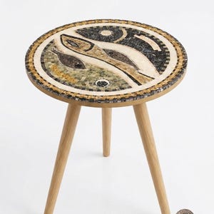 Pode incluir: Uma mesa lateral redonda com um tampo de mosaico apresentando um padrão inspirado em peixes em tons de bege, verde, preto e branco. O tampo da mesa é contornado por uma borda geométrica e apoiado por três pernas de madeira clara em tripé.
