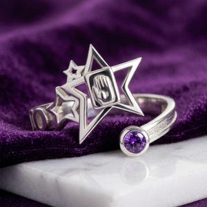 Puede incluir: Un anillo abierto de tono plateado que presenta un motivo de estrella grande con un símbolo de mano en relieve en el centro, acompañado de dos estrellas recortadas más pequeñas. Un extremo de la banda termina en una gema morada facetada.