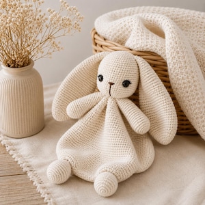 May include: A cream-colored crochet security blanket featuring a stuffed bunny head with long ears, small arms, and two round feet. The toy is crafted with a tight knit texture, featuring dark brown embroidered eyes and a nose on the bunny face.