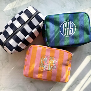 May include: Three rectangular zippered pouches. One features a black and white checkered pattern. Another is blue and green striped with a white circular monogram reading GHS. The third is orange and pink striped with a light blue monogram KGS.