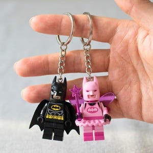 May include: Two keychains featuring plastic minifigures. One is a black Batman character with a cape and yellow emblem. The other is a pink fairy character with wings, a tutu, and a star-tipped wand. Both are attached to silver metal rings.