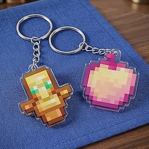 May include: Two acrylic keychains featuring pixelated designs. One keychain is a brown and yellow totem figure with green accents, and the other is a round, pink and yellow fruit-shaped charm. Both include silver metal rings and chains.