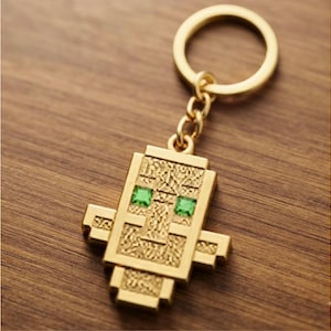Puede incluir: Un llavero de metal dorado con un colgante de personaje pixelado y anguloso, con una superficie texturizada y dos pequeños ojos cuadrados de piedra preciosa de color verde brillante. El colgante está unido a una anilla metálica.