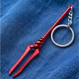May include: A red metal keychain featuring a long, slender, spear-like charm with a spiraled handle and a two-pronged tip. The charm is attached to a standard silver-colored circular metal split ring by a short silver-colored metal chain link.