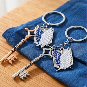 Op de afbeelding: Twee metalen sleutelhangers in de vorm van een sleutel met een schildvormig embleem. De ene sleutel is roségoud en de andere is zilverkleurig. Elk embleem toont witte vleugels met blauwe accenten op een schild. De sleutels hebben Japanse tekens gegraveerd.