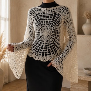 May include: A cream-colored crochet poncho featuring an intricate spiderweb-inspired pattern that radiates from the center. The garment has long, wide bell sleeves and a scalloped hemline, worn over a solid black long-sleeved base layer.