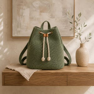 Può includere: Uno zaino all'uncinetto color verde salvia con chiusura a coulisse dotata di spesse corde color crema, un manico superiore e due spallacci. La borsa presenta una base strutturata e un motivo a maglia testurizzato su tutta la superficie.