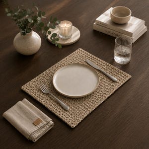 Puede incluir: Un mantel individual rectangular de ganchillo en color beige con un tejido texturizado, centrado con un plato de cerámica redondo, un tenedor de plata a la izquierda y un cuchillo de plata a la derecha. Una servilleta de lino beige doblada.