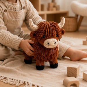 Pode incluir: Um peluche de vaca Highland feito em croché castanho com uma juba de fios desgrenhados, chifres cor de creme, um focinho claro e cascos pretos. O brinquedo apresenta olhos escuros e um corpo texturizado feito de fios com pontos detalhados.