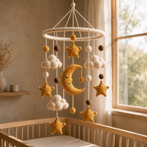 May include: A crochet baby mobile featuring a central yellow crescent moon with a stitched face, surrounded by hanging cream-coloured clouds, yellow stars, and small brown and cream spheres suspended from a circular white ring with white cords.