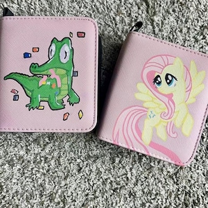 May include: Two square pink wallets with zip closures. The left wallet features a green crocodile character with colorful rectangular shapes. The right wallet displays a yellow pony character with a pink mane, wings, and a blue butterfly cutie mark.