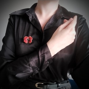May include: A vibrant red poppy flower brooch with dark centre details pinned to a black long-sleeved button-down shirt. The shirt features a classic collar, buttoned cuffs, and a black belt with a gold-toned buckle worn at the waist.